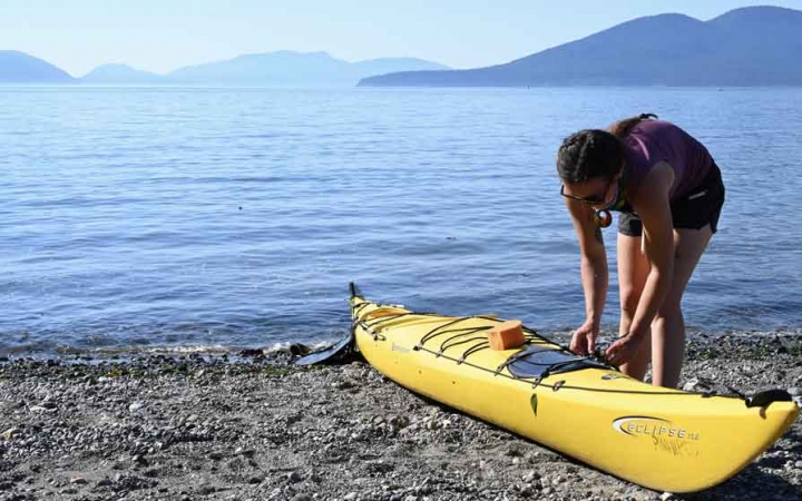 a person fixes something to a yellow kayak on the shore of a very blue lake. there are mountains in the background.